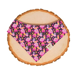 May include: A black dog bandana with a repeating pattern of pink UFOs, planets, astronauts, and moons. The bandana is made of fabric and is displayed on a wooden surface. The text "sitty goose" is visible on the UFOs.