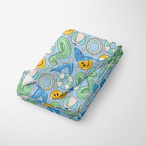 Könnte beinhalten: Ein blaues Strandtuch mit einem sich wiederholenden Muster aus grünen Dinosauriern, blauen Haien, gelben Smiley-Gesichtern, blauen und weißen Rettungsringen und blauen und weißen Strandbällen.