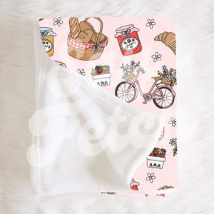 Puede incluir: Una manta de forro polar rosa y blanca con un patrón repetido de una bicicleta, tarros de mermelada, pan y cestas de bayas. La manta está doblada por la mitad, mostrando el forro polar blanco en un lado y el forro polar rosa estampado en el otro.