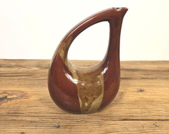May include: A teardrop-shaped ceramic pitcher with a large handle. The pitcher is a deep reddish-brown with a vertical beige stripe. The handle curves gracefully from the base to the spout. The pitcher is on a wooden surface.