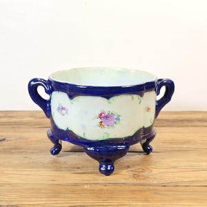 May include: A deep blue ceramic bowl with two handles and three feet. The exterior features a central white panel decorated with a hand-painted floral motif in shades of purple, pink, and green, set against a glossy, dark cobalt blue finish.