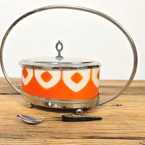 May include: A vintage sugar bowl with a lid and a small spoon. The bowl is orange and white with a geometric pattern, set in a silver-toned metal holder with a handle. The spoon has a black handle.