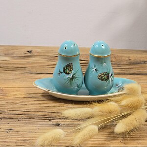 May include: A vintage salt and pepper shaker set. The set includes two light blue shakers and a matching oval tray. Each shaker features a gold rim and a painted pinecone design. The set is displayed on a wooden surface.