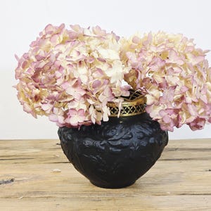 May include: A black, patterned vase displays dried hydrangeas. The flowers are a blend of pale pink and cream, with a gold detail at the top of the vase. The vase rests on a rustic wooden surface.