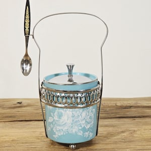 May include: A vintage sugar bowl with a light blue floral design, silver trim, and a matching spoon. The bowl has a lid and sits in a decorative metal holder. The spoon has a black handle with gold accents.