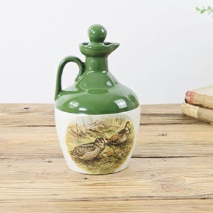 May include: A green and white ceramic jug with a handle and a stopper. The jug features a painted illustration of two birds in a field. The top and handle are a solid green color, while the base is white.