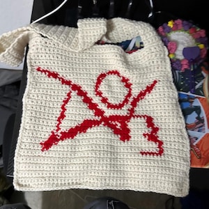 May include: A white crocheted tote bag with a red crocheted symbol of a person with arms outstretched. The bag has a long strap.