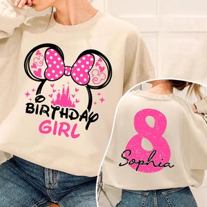 A person wearing a white sweatshirt with a pink and black design, including a large pink number 8 and a Minnie Mouse-inspired bow. The sweatshirt also has the text "BIRTHDAY GIRL" printed on it.