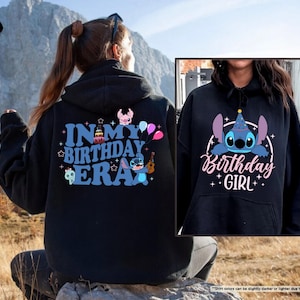 A person wearing a black hoodie with a birthday-themed design, including a character and text, sitting in a field with mountains in the background.