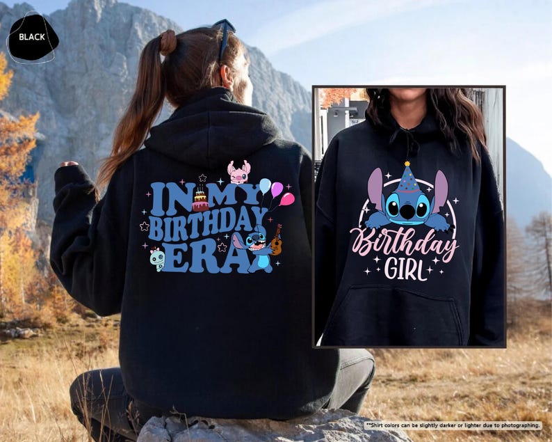 A person wearing a black hoodie with a birthday-themed design, including a character and text, sitting in a field with mountains in the background.
