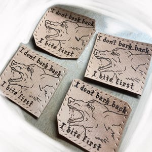 Może przedstawiać: Cztery kwadratowe, jasnobrązowe skórzane naszywki z napisem "I don't bark back, I bite first" i ilustracją ryczącego niedźwiedzia. Naszywki mają szytą obwódkę i są ułożone w kwadrat.