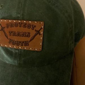 Puede incluir: Gorra de béisbol verde oscuro con un parche de cuero marrón. El parche está cosido a la gorra y presenta las palabras "PROTECT TRANS YOUTH" en mayúsculas, con dos espadas cruzadas.