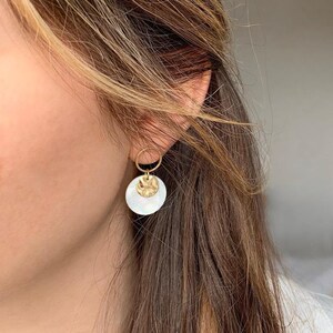 Peut inclure: Boucles d'oreilles dorées à deux niveaux. Le niveau supérieur est un petit disque doré texturé, et le niveau inférieur est un disque blanc nacré plus grand. Les boucles d'oreilles sont suspendues à un petit cercle doré. Les boucles d'oreilles sont portées.