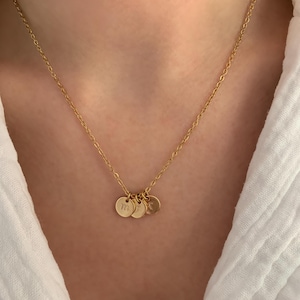 Op de afbeelding: Een gouden ketting met drie kleine ronde bedels. Elke bedel heeft een andere letter gegraveerd.