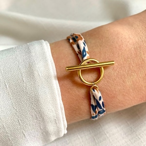 Peut inclure: Bracelet à fermoir en or avec un bracelet en tissu à motifs. Le bracelet présente des motifs floraux bleus et bruns sur fond blanc. Le bracelet est porté sur un poignet, avec une manche de chemise blanche en arrière-plan.