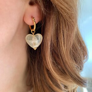 Peut inclure: Boucles d'oreilles créoles dorées avec un pendentif en forme de cœur blanc. Le pendentif a un motif en spirale.