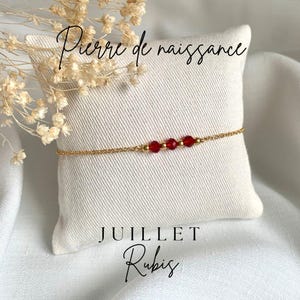 Puede incluir: Un brazalete de cadena dorada con cuentas de rubí rojo. El brazalete está sobre un cojín blanco con el texto "Pierre de naissance" y "JUILLET Rubis".