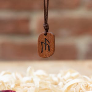May include: A brown wooden pendant with a dark engraved symbol, suspended from a brown cord. The pendant has a smooth, rounded rectangular shape with visible wood grain texture, and the black symbol is centered on the front surface of the wood.