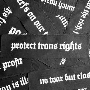 May include: Black fabric patches with white text in a gothic font. The patches read "protect trans rights", "no war but class", "profit", "on is ill", and "erfs on our t".