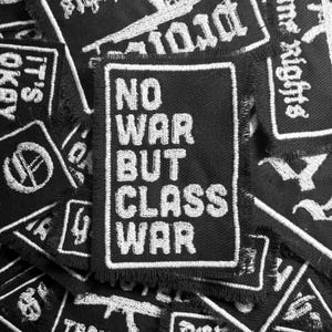 May include: A black and white embroidered patch with the text "NO WAR BUT CLASS WAR" in white lettering.