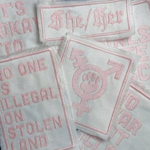 May include: A collection of white fabric patches with pink embroidered text and symbols. The patches include the text "It's okay to", "No one is illegal on stolen land", "She/Her", and a transgender symbol with a fist.