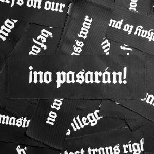 May include: A black fabric patch with white text that reads "ino pasarán!" The patch is surrounded by other patches with different text, all in a similar style.