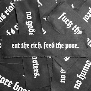 May include: A collection of black fabric patches with white text. The patches have different slogans, including "eat the rich, feed the poor."