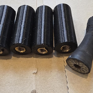 May include: Four black cylindrical components with visible threaded metal inserts at the base, arranged in a row, alongside a single black tapered handle-like object with a textured surface and a circular opening at the end.