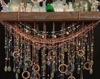 May include: A rustic wooden shelf with a copper wire and bead chandelier hanging below. The chandelier features a variety of blue, green, and clear glass beads, along with small glass bottles and birds.