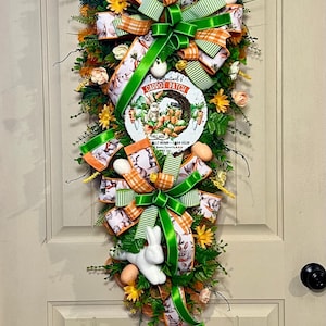 May include: A decorative Easter wreath with a horseshoe-shaped sign that reads "Carrot Patch". The wreath features green and orange ribbons, faux eggs, a white bunny figurine, and floral accents. The wreath is designed to hang on a door.