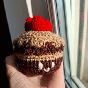 Può includere: Un cupcake all'uncinetto con una ciliegia rossa sopra. Il cupcake ha una combinazione di colori marrone e beige, con un volto sorridente ricamato sul fondo. L'oggetto fatto a mano è tenuto in una mano.