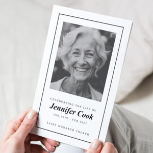 Puede incluir: Una tarjeta conmemorativa en blanco y negro sostenida por una persona. La tarjeta presenta una fotografía de una mujer sonriente, con el texto "Celebrando la vida de Jennifer Cook" y las fechas "Ene 1970 - Feb 2027". El nombre de la iglesia también está impreso en la tarjeta.