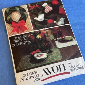 May include: A vintage tapestry pattern collection booklet featuring Christmas-themed designs. The cover displays a braided wreath with a red bow, a Christmas tree with ornaments, and placemats. The booklet is designed exclusively for Avon.