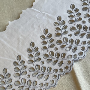 May include: Close-up of a white eyelet lace trim with a scalloped edge. The lace features a repeating pattern of stylized leaves in a light gray color, creating an elegant and delicate design. Ideal for sewing and craft projects.