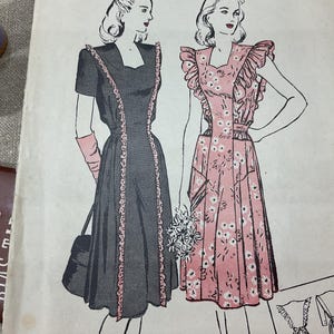 May include: Vintage sewing pattern featuring two dress designs. One dress is dark gray with pink trim and a matching purse. The other is pink with a floral pattern and ruffled sleeves. The pattern is on aged paper with spools of thread in the background.