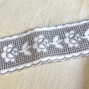 May include: Close-up of a decorative lace trim featuring a floral design. The lace is white with a gray floral pattern and a scalloped edge. The trim is placed on a beige linen fabric.