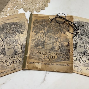 May include: Three vintage agricultural almanacs from 1931, 1945, and 1953, with black-rimmed glasses resting on the center book. The covers feature illustrations of women and text, including "Agricultural Almanac" and the year.