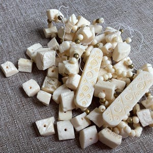 May include: Ivory-colored beads and carved bone pieces scattered on a textured surface. The collection includes small cubes, rectangular pieces with etched designs, and strung beads with gold-colored accents. A neutral, natural aesthetic.
