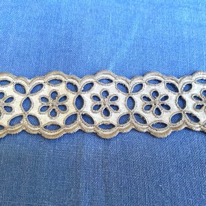 May include: Close-up of a cream-colored lace trim with a floral design. The trim has a scalloped edge and is set against a blue fabric background. The lace features openwork details and a delicate, textured appearance.
