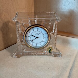 May include: A clear crystal clock with a round face featuring Roman numerals and the words "Waterford Crystal." The clock has a gold-colored bezel and a decorative crystal frame. The clock is sitting on a white surface.