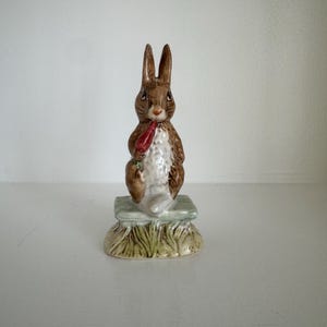 May include: A brown ceramic figurine of a rabbit sitting on a green base. The rabbit is holding a red carrot in its paws.