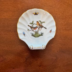 May include: A white, shell-shaped dish with a gold rim. The dish features a detailed illustration of two birds perched on a branch with green leaves. A butterfly and insects are also depicted. The dish is likely porcelain or ceramic.