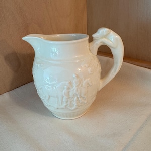 May include: Cream-colored ceramic pitcher with a dog-shaped handle. The pitcher features a detailed relief design around the body, depicting figures and animals. The spout is curved, and the overall design is classic and elegant.