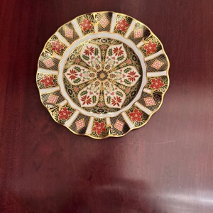 May include: Decorative plate with a scalloped edge. The plate features a central floral design in white, red, and green, surrounded by a black and gold geometric pattern. The rim is also decorated with red floral motifs.