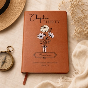 May include: A brown notebook featuring the text Chapter THIRTY, a floral illustration, the name Hayden in a decorative frame, and the text EVERY CHAPTER BRINGS NEW GROWTH. The cover has a smooth texture and a matching brown ribbon bookmark.