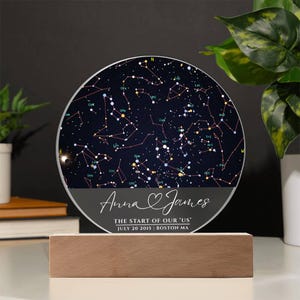 Puede incluir: Un mapa del cielo nocturno personalizado, exhibido en un panel acrílico circular transparente. El mapa presenta constelaciones y estrellas, con los nombres "Anna & James" y la fecha "July 20 2015 | Boston MA" impresos debajo. El panel descansa sobre una base de madera.