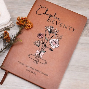 May include: A brown leather journal with the words "Chapter Seventy" and a floral bouquet design. The journal includes the name "Mary" and the quote "Every chapter brings new growth."