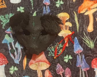 May include: A black mask with fluffy ears and a red string, resting on a dark fabric patterned with colorful mushrooms and foliage. The mushrooms are in shades of red, orange, yellow, and blue, creating a whimsical design.