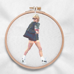 May include: A cross-stitch embroidery hoop with a colorful image of a person wearing a blue jacket, white shorts, and white sneakers. The person is walking and has short blonde hair.