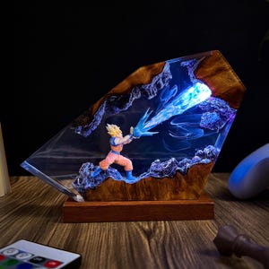 May include: A decorative resin lamp featuring a figure in an action pose, emitting a blue energy blast. The lamp has a wooden base and frame, with a clear resin front. The figure is wearing an orange and blue outfit. The lamp is lit with blue and white lights.